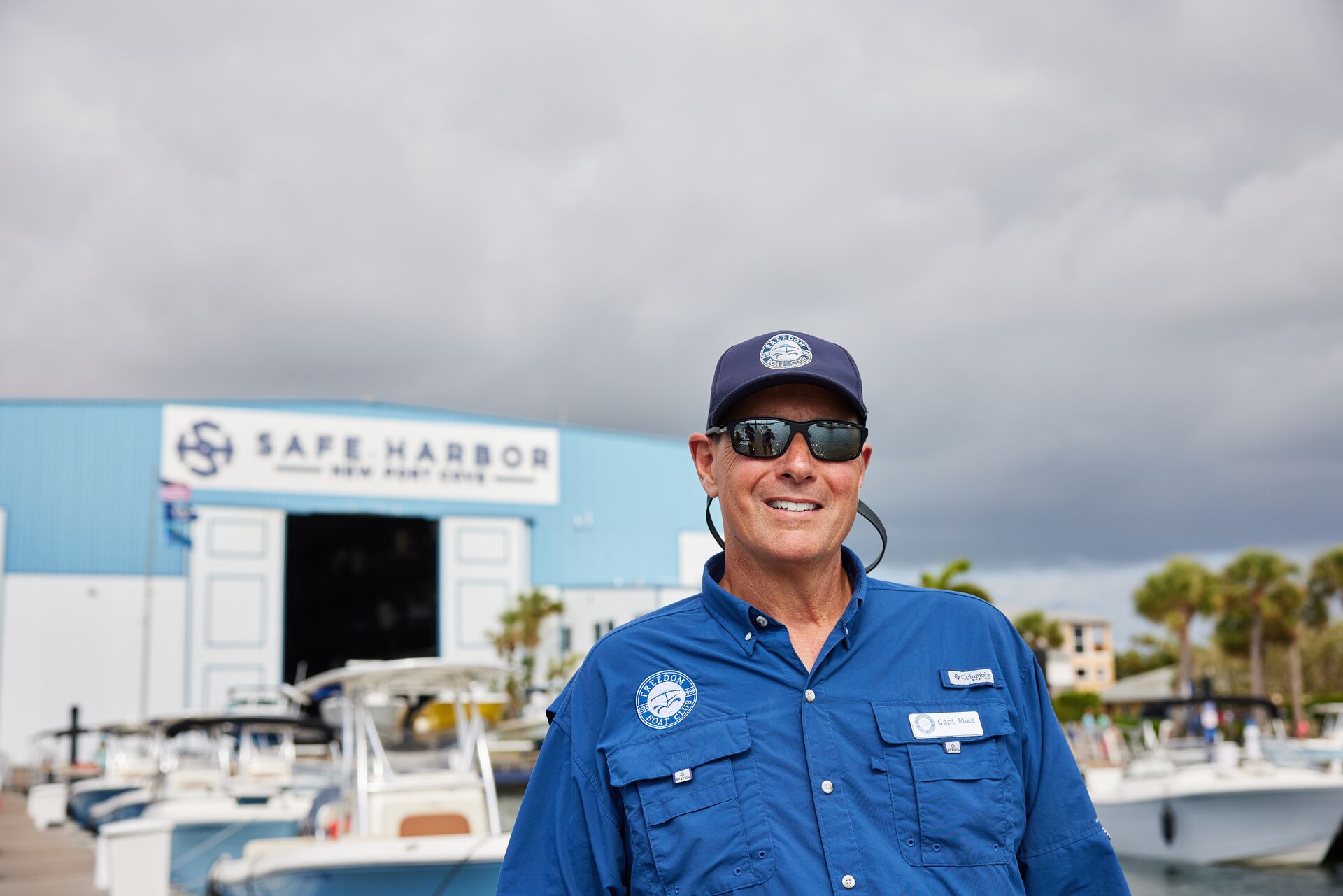 Man smiles while wearing sunglasses at a boat harbor.